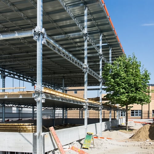 The image shows a construction site with a partially built structure featuring metal trusses and concrete base There is a tree on the right and a building in the background-1